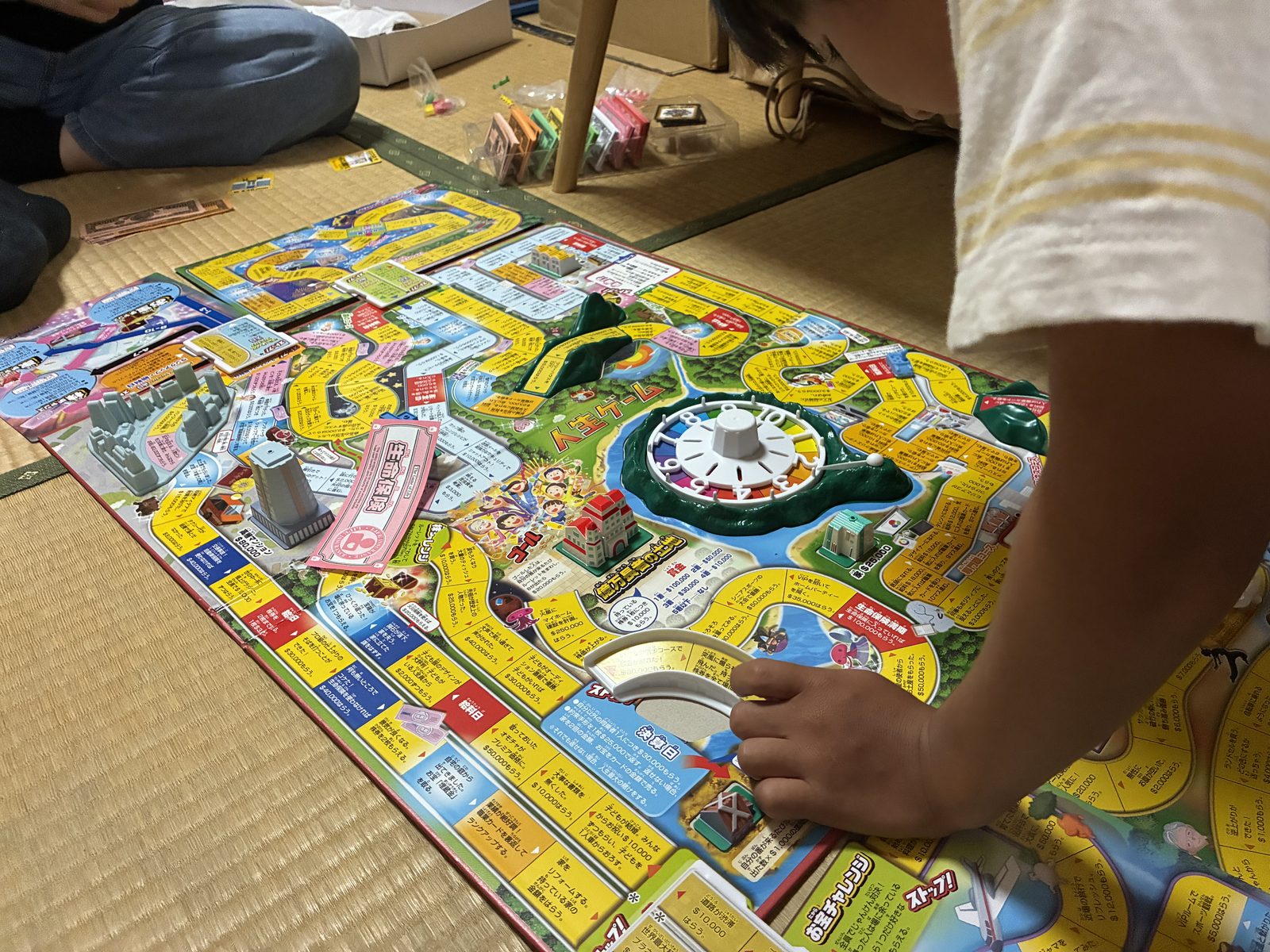 畳の部屋に広げた人生ゲームの盤を、子どもと大人が囲んでいる様子